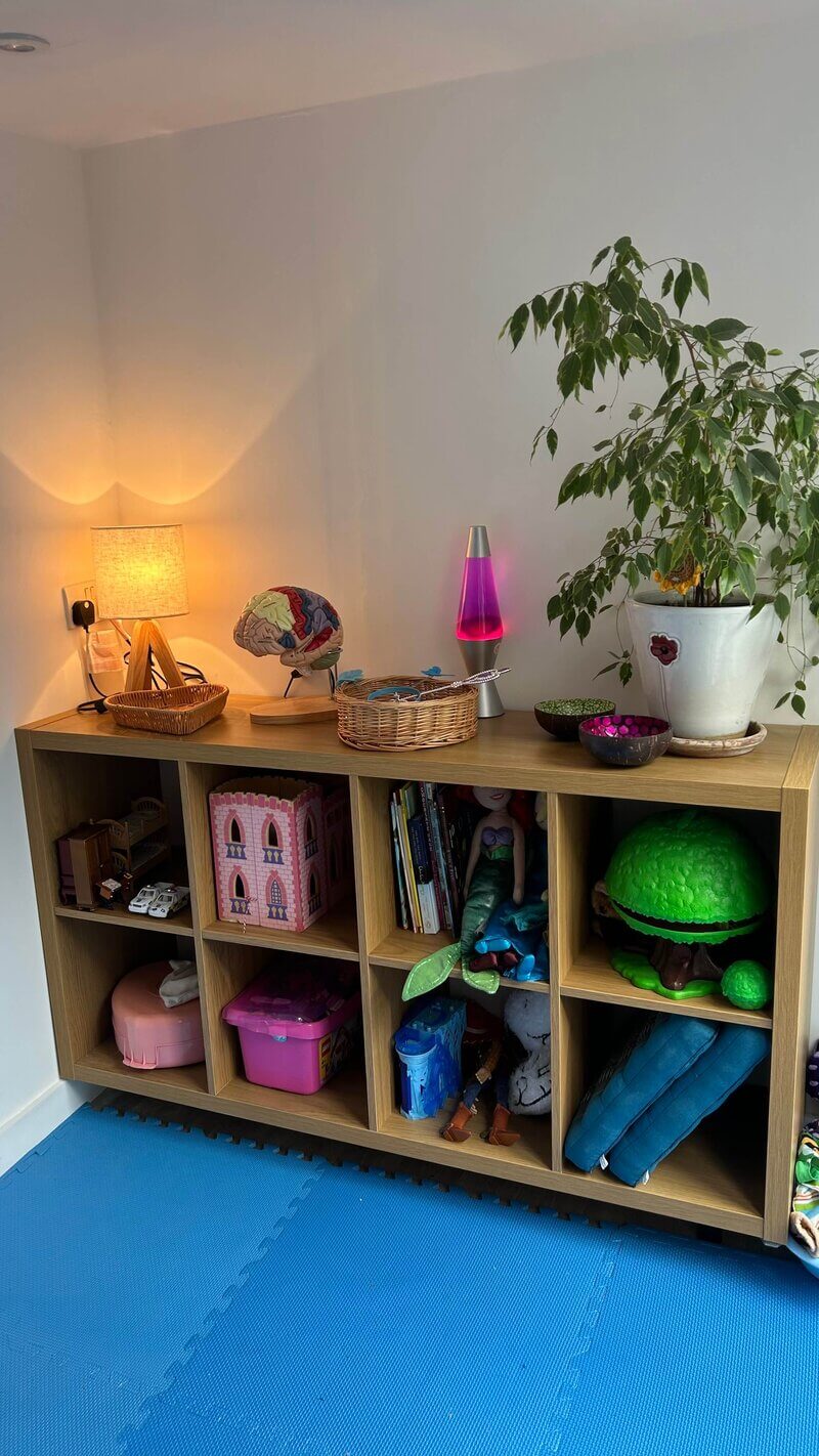 Interior of 'The Sunflower House', the play therapy room of Turn To The Sun Play Therapy. The image shows a display shelf with 4 main square compartments filled with children's play things. On top of the shelf is a lamp, a lava lamp, a plant pot, and various containers.