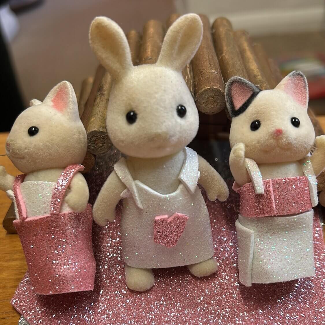 Three small toy figures — a bunny and two cats — dressed in glittery clothes, standing together on pink and silver glitter paper.