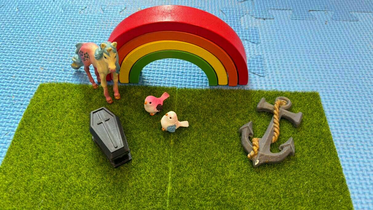 A symbolic arrangement with a rainbow, a small coffin, two birds, a horse, and an anchor placed on grass