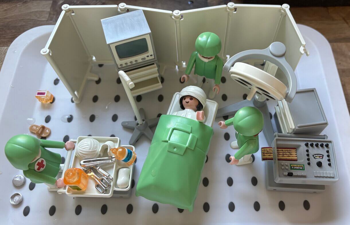 Playmobil figure lying in a toy hospital bed surrounded by medical equipment and doctors