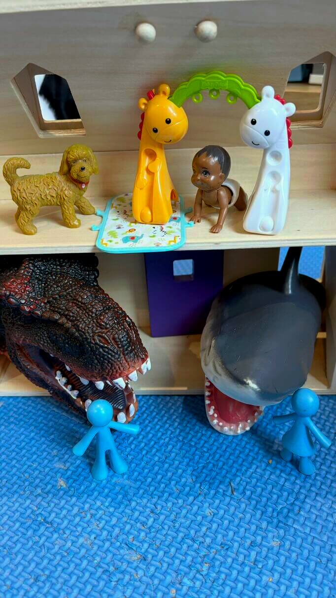 Two toy scenes stacked vertically — one with a baby and animals above, and another with a dinosaur and shark confronting small blue figures below
