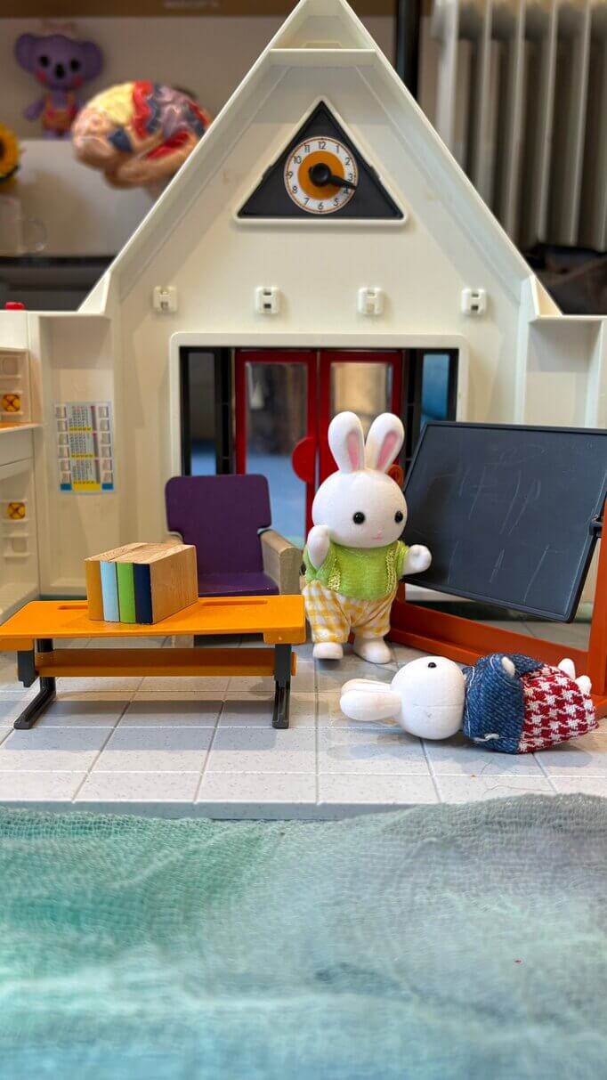 A toy rabbit figure lying on the floor of a school playset, while another rabbit stands nearby near a chalkboard