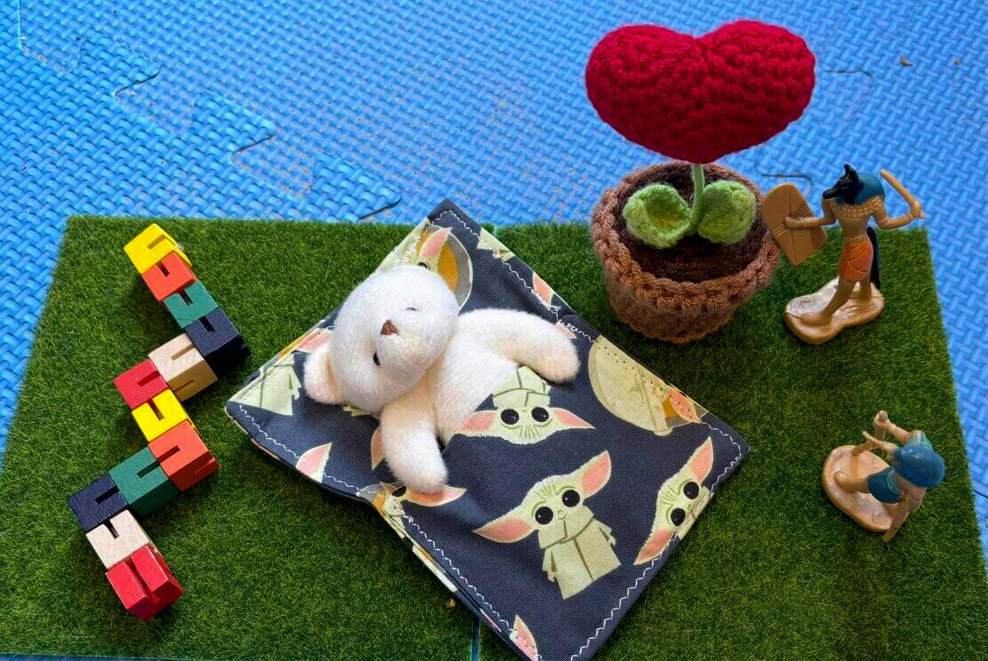 A soft toy bear tucked under a blanket beside symbolic and mythological figures, a red heart plant, and puzzle pieces